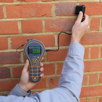 Abgebildet ist das Feuchtigkeitsmessgerät MMS3 der Firma PROTIMETER, das in einer Hand gehalten wird. In der anderen Hand befinden sich die Eindrückelektroden, die zur Baufeuchtemessung in Prozent eingesetzt werden. Die Messung erfolgt an einer deutlich e